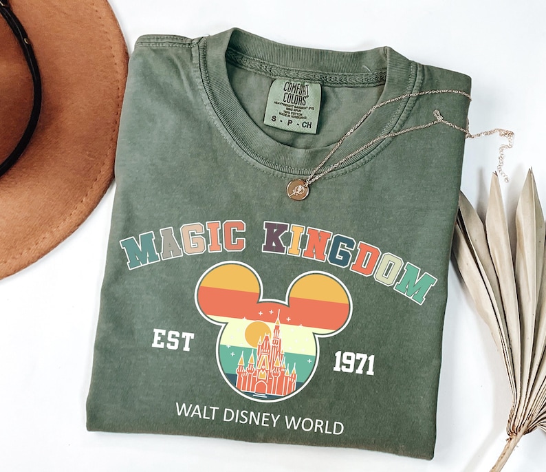 K&ouml;nnte beinhalten: Ein olivgr&uuml;nes T-Shirt mit einer farbenfrohen Grafik mit den Worten "MAGIC KINGDOM" und einer Mickey Mouse-Silhouette mit einem Schlossdesign. Das Shirt enth&auml;lt auch den Text "EST 1971" und "WALT DISNEY WORLD". Eine goldene Halskette liegt auf dem Shirt.