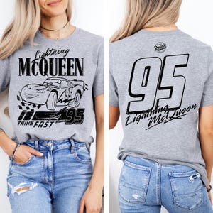 May include: Gray t-shirt featuring Lightning McQueen from the Cars movie. The front graphic includes the character's image, the text "Lightning McQueen," and "95." The back displays a large "95" and "Lightning McQueen" text. The shirt is worn with blue jeans.
