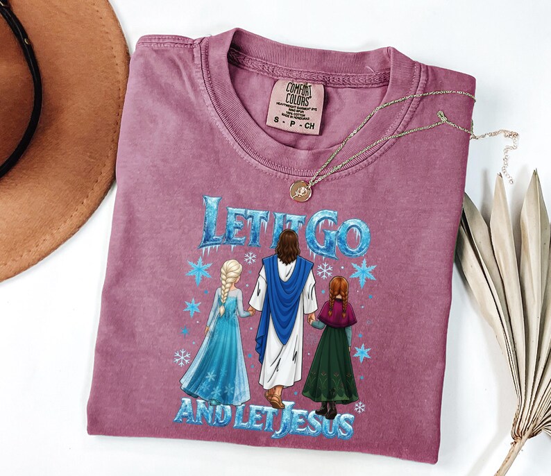 May include: A mauve Comfort Colors t-shirt with the text "LET IT GO AND LET JESUS" in blue. The graphic depicts Jesus holding hands with two characters from the movie Frozen. A gold necklace rests on the shirt.