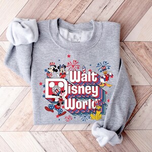 K&ouml;nnte beinhalten: Graues Sweatshirt mit farbenfroher Grafik mit der Aufschrift "Walt Disney World". Das Design zeigt Micky Maus, Minnie Maus, Donald Duck und Pluto, mit Feuerwerk und Sternen.