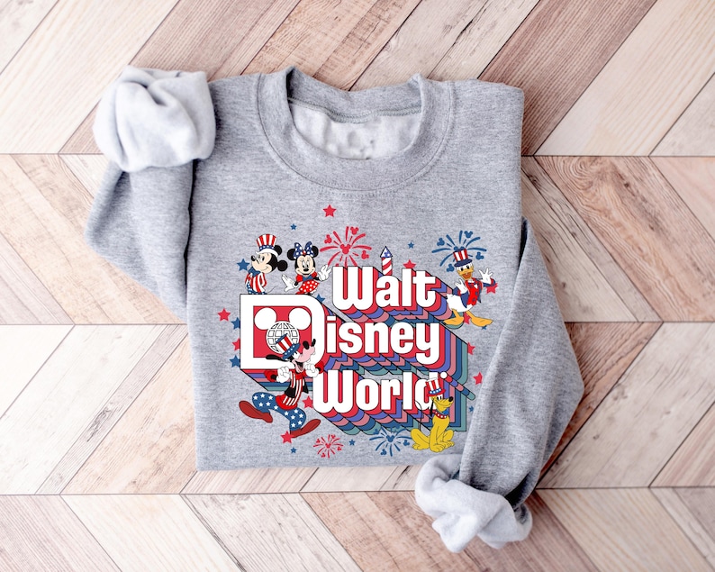 K&ouml;nnte beinhalten: Graues Sweatshirt mit farbenfroher Grafik mit der Aufschrift "Walt Disney World". Das Design zeigt Micky Maus, Minnie Maus, Donald Duck und Pluto, mit Feuerwerk und Sternen.