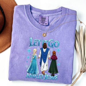 May include: A lilac Comfort Colors t-shirt with the text "LET IT GO AND LET JESUS" in blue and white. The graphic shows Jesus with two animated characters. A gold necklace with a pendant is draped across the shirt.