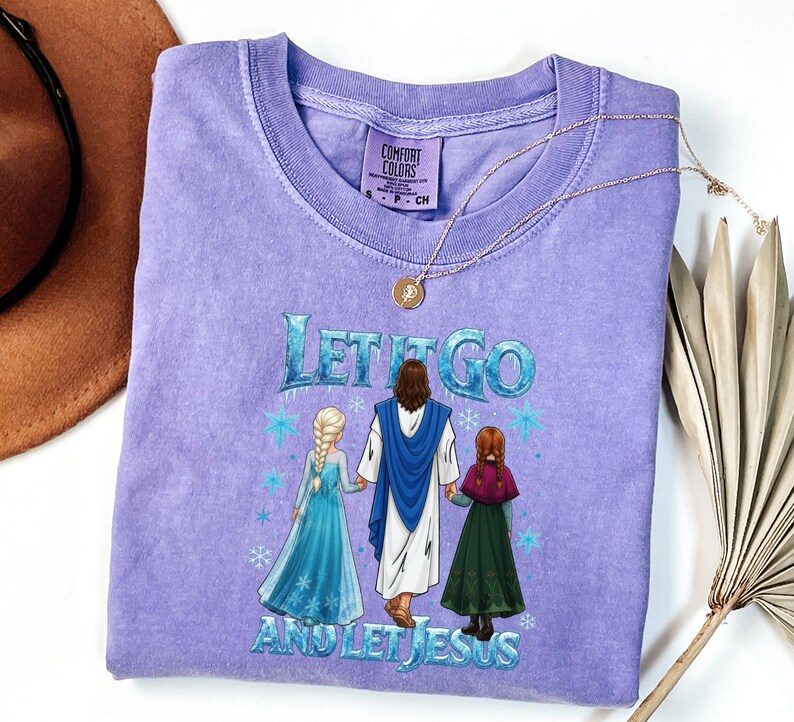 May include: A lilac Comfort Colors t-shirt with the text "LET IT GO AND LET JESUS" in blue and white. The graphic shows Jesus with two animated characters. A gold necklace with a pendant is draped across the shirt.