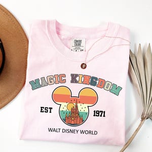 K&ouml;nnte beinhalten: Ein hellrosa T-Shirt mit dem Schriftzug "MAGIC KINGDOM" in einem Retro-Schriftstil und einer Mickey-Mouse-Kopf-Grafik mit einem Schloss darin. Auf dem Shirt steht au&szlig;erdem "EST 1971" und "WALT DISNEY WORLD". Eine goldene Halskette liegt auf dem Shirt.