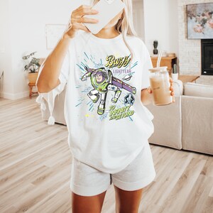 May include: White t-shirt featuring a graphic of Buzz Lightyear from Toy Story. The design includes the text "Buzz Lightyear" and "To Infinity and Beyond" in a colorful, cartoon style. The shirt is a casual, oversized fit.