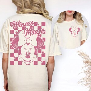 May include: Cream-colored t-shirt featuring a pink Minnie Mouse graphic. The back of the shirt displays a large Minnie Mouse design with the words "Minnie Mouse" above the character. The front of the shirt has a smaller Minnie Mouse graphic on the chest.