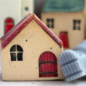 May include: A small ceramic house figurine with a cream-colored facade, a red gabled roof, and a red arched door. The structure features a single arched window with a black frame. A light blue object with a ridged texture is positioned to the right.