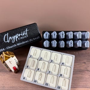 May include: A collection of church-themed craft supplies. Includes a black box with "Clayprint Studio" text, a small ceramic church model, and a set of stamps with various church window designs. The stamps are arranged in a black holder and a gray tray.
