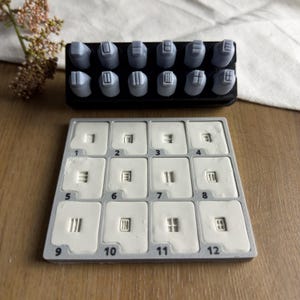 Puede incluir: Un juego de doce sellos grises con varios patrones de líneas geométricas en sus caras, guardados en una bandeja rectangular negra. Debajo de los sellos hay un molde cuadrado gris con doce compartimentos numerados, cada uno con una impresión de arcilla blanca.