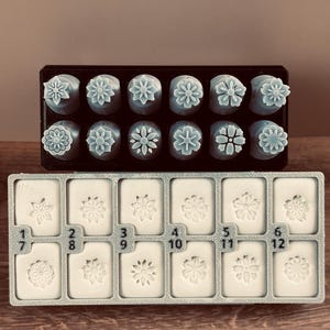 May include: A set of twelve flower-shaped soap stamps and twelve flower-shaped soap molds. The stamps are arranged in a black tray, and the molds are in a gray tray. Each stamp and mold has a unique floral design and is numbered.