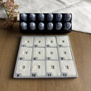 Puede incluir: Un juego de doce sellos de metal gris dispuestos en un soporte negro, colocados sobre una bandeja cuadrada gris que contiene doce impresiones de arcilla blanca. Cada impresión corresponde a un sello numerado del uno al doce con patrones geométricos.