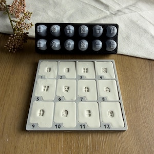 Puede incluir: Un conjunto de doce sellos grises con varios diseños de ventanas y puertas, junto con una bandeja cuadrada gris que contiene doce compartimentos numerados rellenos con impresiones de arcilla blanca de cada diseño, numerados del uno al doce.