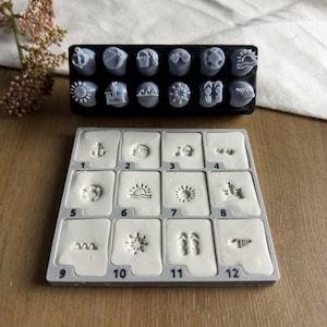 May include: A set of twelve grey rubber stamps featuring summer-themed icons including an anchor, beach ball, bucket, sunglasses, life preserver, sun, waves, sandcastle, starfish, flip-flops, and seashell, paired with a white clay impression tray.