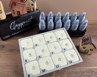 May include: A collection of clay art tools and miniature ceramic houses. The tools include various shaped stamps and nozzles. The text "Clayprint Studio" is visible on a black base. The stamps are numbered 1-12.
