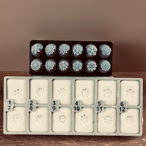 以下が含まれることがあります： 2つの花の形をしたクッキー型スタンプのセット。上のセットは黒いトレイに、下のセットは1〜12の番号が付いた薄い灰色のトレイに入っています。スタンプは水色と白です。