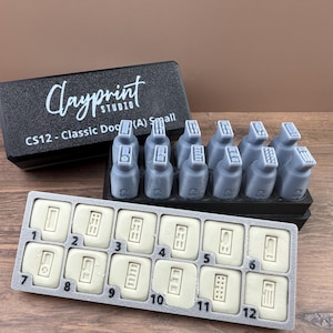 May include: A set of gray clay stamps with a black box that reads "Clayprint Studio" and "CS12 - Classic Dog (A) Small." The stamps are arranged in a black tray and a gray tray with numbered rectangular molds.