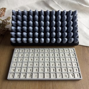 May include: A set of gray metal stamps arranged in a grid, paired with a rectangular gray silicone mold featuring recessed letters, numbers, and symbols including A-Z, a-z, 0-9, #, @, !, and punctuation marks in a clear, sans-serif font.