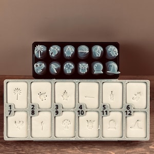 May include: Two trays of crafting stamps. The top tray holds twelve light blue stamps with various designs, including a palm tree, mushroom, and owl. The bottom tray has twelve white stamps with corresponding designs and numbers.