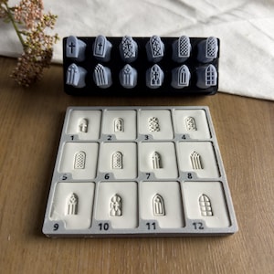 May include: A set of twelve grey stamps featuring various gothic window and cross designs, paired with a matching grey silicone mould tray containing twelve numbered indentations from one to twelve, each corresponding to the specific stamp patterns.