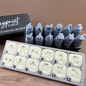 May include: A set of Christmas-themed clay stamps and molds. The set includes 12 molds with festive designs like a snowman, gingerbread man, and Christmas tree. The gray stamps are in a black holder with the text "Clayprint Studio".