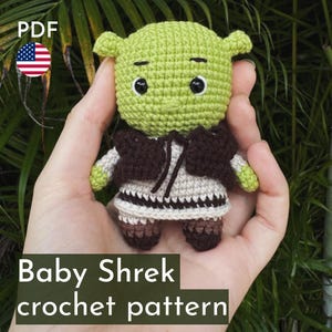 May include: A crochet pattern for a green, brown and beige amigurumi baby Shrek doll. The doll is holding a small brown bag. The pattern is available as a PDF download.