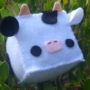Puede incluir: Un juguete de peluche de vaca blanco y negro con una nariz rosa y ojos negros. El juguete tiene forma de cubo y tiene una costura cosida alrededor de los bordes.