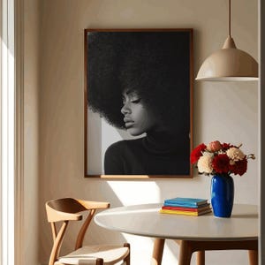 May include: A black and white photograph of a woman with an afro hairstyle wearing a black turtleneck sweater. The photo is framed and hanging on a wall in a room with a wooden table, chairs, and a vase of flowers.
