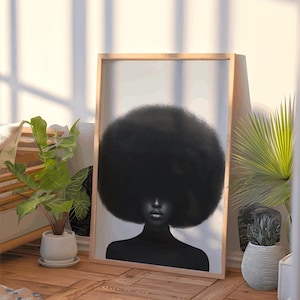 May include: A framed art print featuring a black and white portrait of a person with a large afro. The person is wearing a black turtleneck. The artwork is displayed in a light wooden frame, leaning against a wall. The room has plants and natural light.