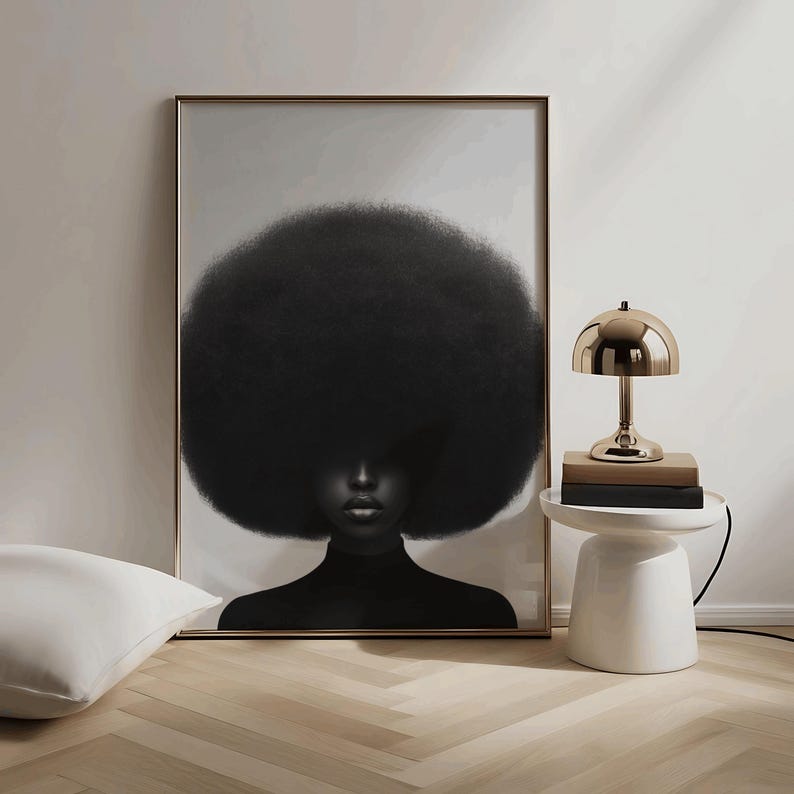 May include: A framed art print featuring a portrait of a person with a large, dark afro. The person is wearing a black turtleneck. A gold-colored lamp sits on a white side table next to the artwork. A white pillow rests on the floor.