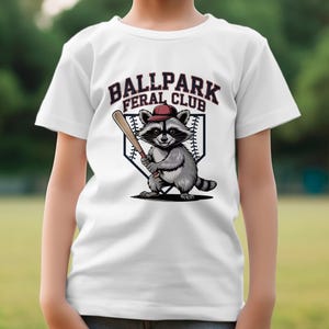 Op de afbeelding: Wit T-shirt met een afbeelding van een wasbeer die een baseballpet draagt en een knuppel vasthoudt. De tekst "Ballpark Feral Club" staat boven de wasbeer.