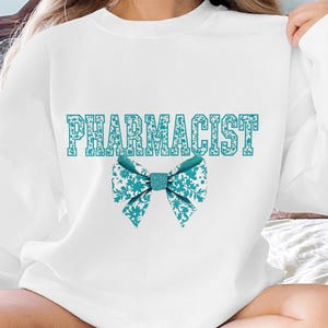 May include: White sweatshirt with the word "PHARMACIST" printed in teal blue letters with a teal blue bow with a white floral pattern.