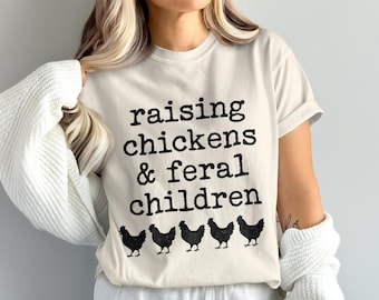 May include: Cream-colored t-shirt with the text "raising chickens & feral children" in black, above a row of five black chicken silhouettes. The shirt has short sleeves and a casual fit.