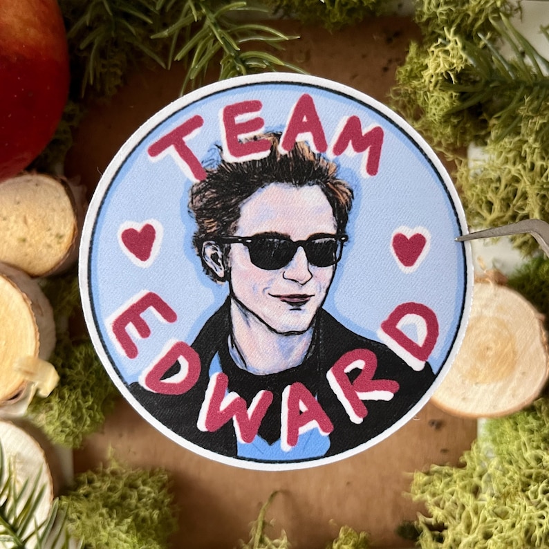 May include: A round sticker with a blue background and white border. The sticker features a cartoon illustration of a man with dark hair and sunglasses. The text "TEAM EDWARD" is written in red letters around the image. Two red hearts are on either side of the image.