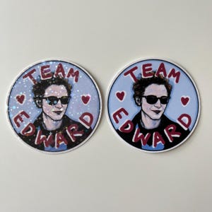 May include: Two round stickers with a blue background and red text. The text on both stickers reads "Team Edward". The stickers feature a drawing of a man with dark hair, wearing sunglasses and a black jacket. The man is smiling. One sticker has a glittery finish.