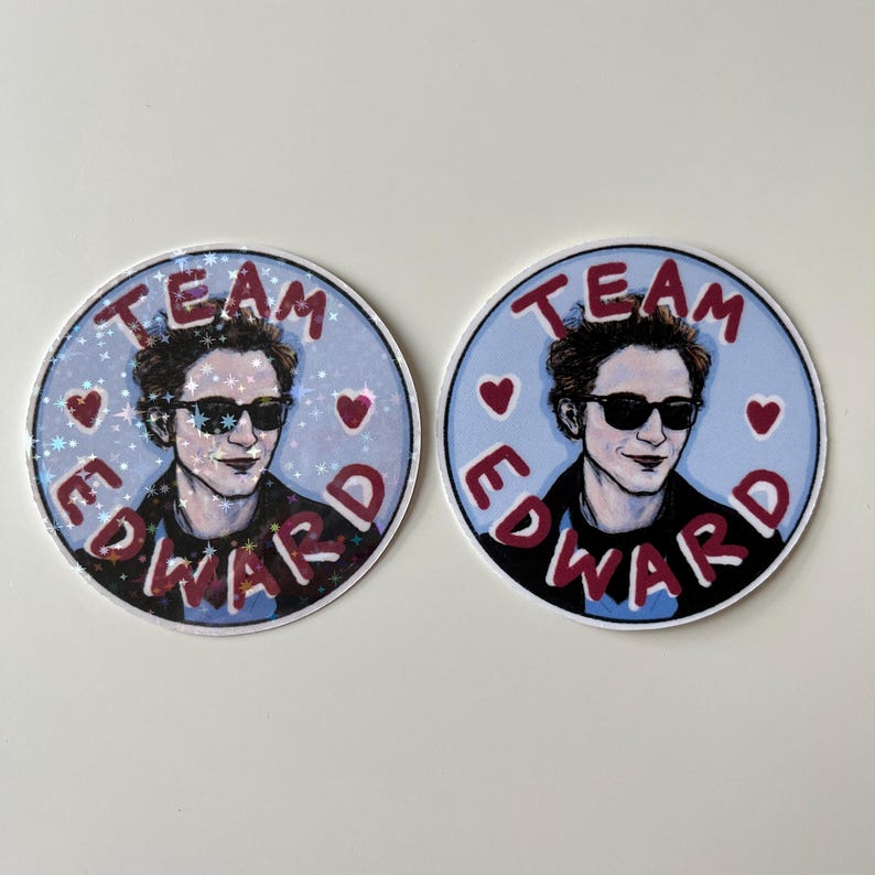 May include: Two round stickers with a blue background and red text. The text on both stickers reads "Team Edward". The stickers feature a drawing of a man with dark hair, wearing sunglasses and a black jacket. The man is smiling. One sticker has a glittery finish.