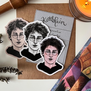 May include: Three Harry Potter stickers with different colored backgrounds. The stickers are black and white illustrations of Harry Potter with his glasses and lightning bolt scar. The stickers are on a brown surface with a business card that says "KettleRain" and "KettleRainArt".