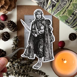 May include: Black and white illustration of a character from a fantasy novel, wearing a cloak and holding a sword. The character is standing on a white background.