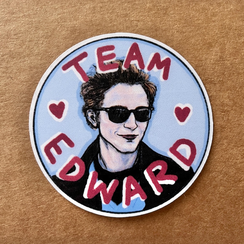 May include: A round sticker with a blue background and a red border. The sticker features a cartoon illustration of Edward Cullen from the Twilight series wearing sunglasses. The text "TEAM EDWARD" is written in red letters around the image.