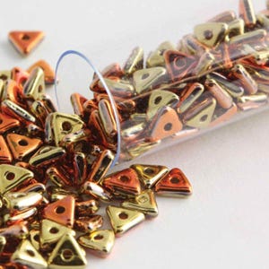 May include: A clear glass tube filled with small, triangular beads. The beads are a mix of gold and copper colors with a shiny, metallic finish.