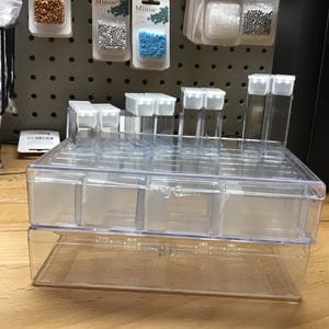 May include: A clear plastic storage box with multiple compartments. The box is stacked on top of another clear plastic storage box. The boxes are ideal for organizing small items like beads, buttons, or jewelry.