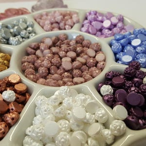 May include: Assortment of colorful, decorative, round, and flower-shaped beads in a white ceramic dish with multiple compartments.