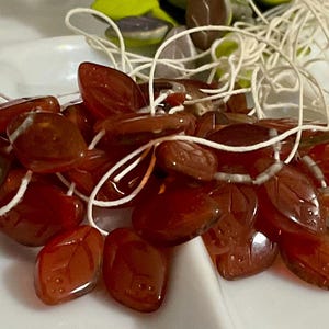 May include: A pile of small, red, glass leaf-shaped beads with a white string running through them. The beads are arranged in a random pattern and are all the same size and shape.