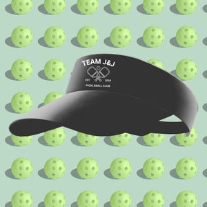 K&ouml;nnte beinhalten: Ein schwarzer Visier mit einem wei&szlig;en Logo, auf dem "TEAM J&J EST 2024 PICKLEBALL CLUB" mit einem gekreuzten Schl&auml;gerdesign steht. Das Visier befindet sich auf einem Hintergrund aus gr&uuml;nen Pickleballs.
