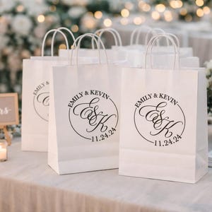 May include: White paper gift bags with handles, each featuring a black circular monogram logo with the text Emily & Kevin and the date 11.24.24 printed on the front. The bags are arranged in a group, displaying the same custom wedding design.