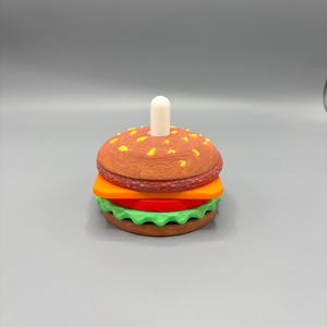 May include: A whimsical burger-shaped object with a brown bun, orange cheese, a red tomato slice, and green lettuce. A white peg is inserted in the top bun. The burger is a playful, colorful design.