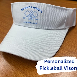 K&ouml;nnte beinhalten: Wei&szlig;er Visier mit blauer Stickerei, auf der "Maggie & Robbie's Est 1994 Pickleball Club" steht, und einer Grafik mit zwei Pickleball-Schl&auml;gern und einem Ball.