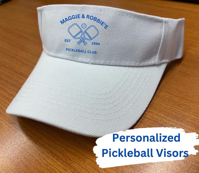K&ouml;nnte beinhalten: Wei&szlig;er Visier mit blauer Stickerei, auf der "Maggie & Robbie's Est 1994 Pickleball Club" steht, und einer Grafik mit zwei Pickleball-Schl&auml;gern und einem Ball.