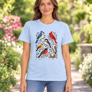 May include: A light blue t-shirt featuring a graphic print of various birds including a blue jay, cardinal, and woodpecker among green leaves and red berries. The text "BACKYARD BIRDS" is printed in small black letters below the bird illustration.