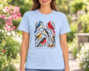May include: A light blue t-shirt featuring a graphic print of various birds including a blue jay, cardinal, and woodpecker among green leaves and red berries. The text "BACKYARD BIRDS" is printed in small black letters below the bird illustration.