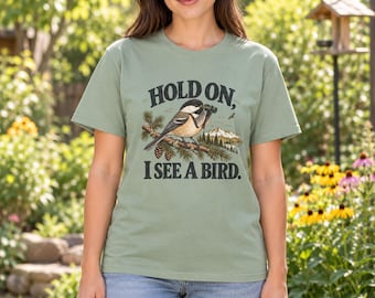 May include: A sage green short-sleeve t-shirt featuring a graphic of a chickadee bird holding binoculars while perched on a pine branch. The text "HOLD ON, I SEE A BIRD." is printed in dark lettering above and below the bird illustration.